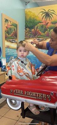 Toddler in Smock getting haircut while sitting in a red car.