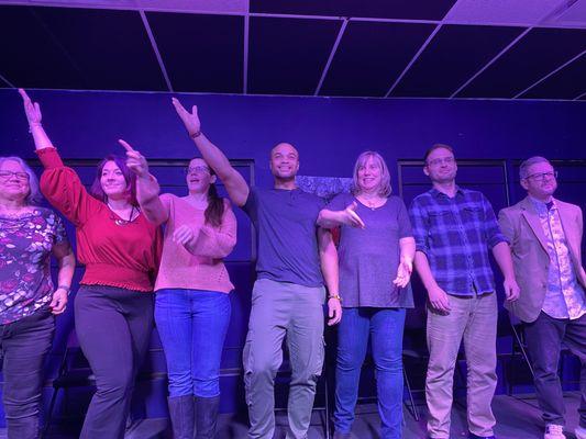 Seven actors onstage, smiling and gesturing to the audience.