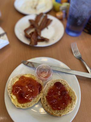 English Muffin with butter and homemade strawberry jam with a side of bacon in the background.