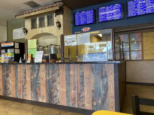 Order counter. Menu is above right. Taped to the plastic on the counter, the yellow sign lists proteins. The green sign lists options.