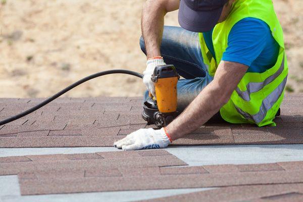 Roofer builder worker with nail gun installing asphalt shingles