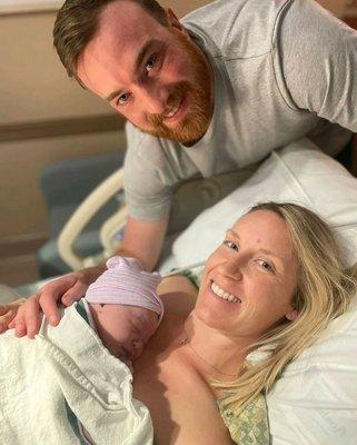 White man with brown hair smiling along with white woman with blonde hair smiling holding her newborn baby on her chest in a hospital bed.