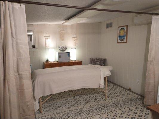 Photo of open curtains and massage table, arm chair, dresser, lamps, towel warmer, and wall decor.