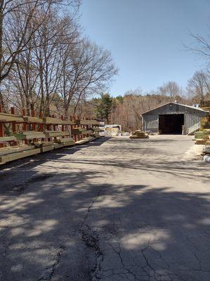 A lumberyard at Builders FirstSource displaying wooden planks and beams stacked on racks outdoors, with a storage shed in the...