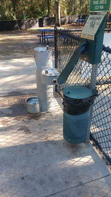 Poop bag dispenser, trash can and water fountain just inside gate