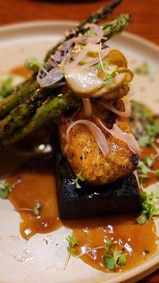 Salmon served on a wood plank served with grilled asparagus and pickled onions, pickles and micro greens .