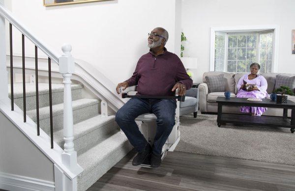 man riding stair lift while wife watches in living room in Greenville, SC