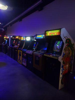 Roughly 12 arcade machines lined up against a wall with dim purple lighting.