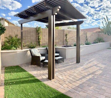 Pavers, freestanding pergola, retaining wall with decorative travertine cap, stucco finish, plants inside raised planter, synthetic grass.