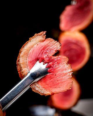 A perfectly cooked slice of Picanha, held by a pair of tongs, ready to be enjoyed.