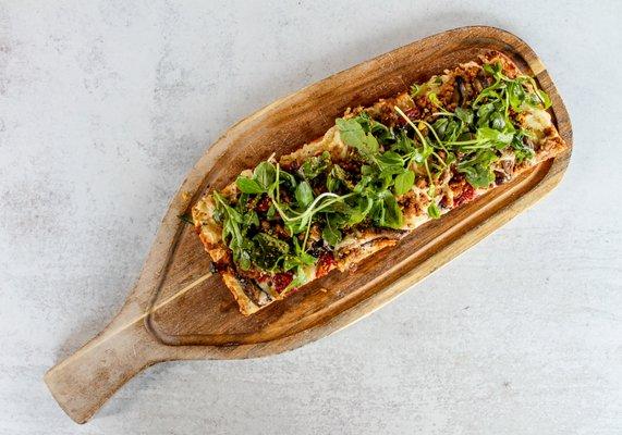 Indulge with our Flatbread with creamy goat cheese, garlic, oak-fired oyster mushrooms, sausage, dried tomato, pistachio, and fresh greens.