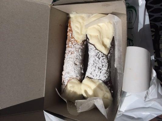 A large cannoli and a large chocolate cannoli, the cream oozing in the sunlight.