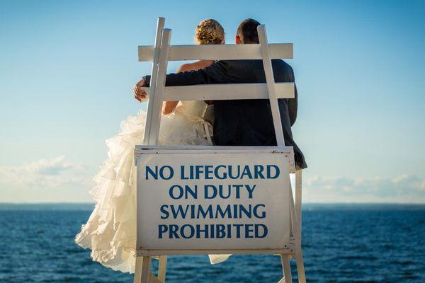 Bride and groom sitting on lifeguard stand looking at ocean