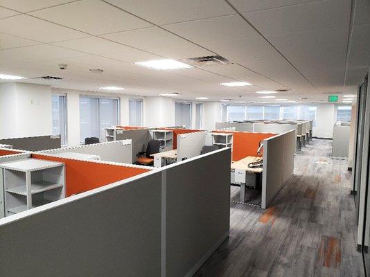 A modern office space featuring a grid of cubicles with gray walls and bright orange accent panels...