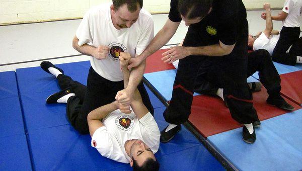 Two men in martial arts uniforms practice techniques on a padded mat. One man lies on the mat holding the other's wrist, while an instructor