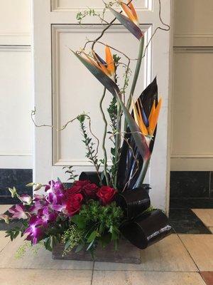 A modern flare of orchids , birds of paradise. And roses , accented with curly willow and ty  leaves designed in a wooden crate .