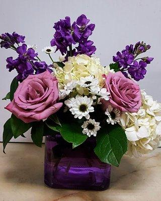 A sweet arrangement of lavender roses and stock, white hydrangea and poms in a purple glass cube