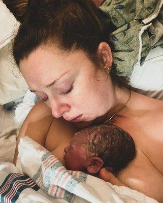 White woman with brown hair pulled up in a bun holding newborn baby against her chest in a hospital bed.