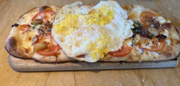 Brunch Flatbread covered with cheese, bacon, crispy potatoes, caramelized onion, tomato, and Parmesan cheese. Topped with a fried egg.