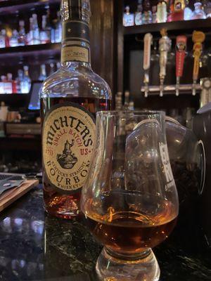 Michters bourbon bottle with a glass (with bourbon). Bottles of bourbon are in the background.