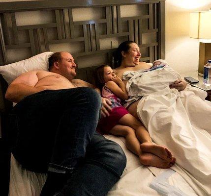 White man shirtless laying next to his older daughter and white laboring woman holding her newborn on her chest in a large queen sized bed.