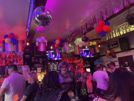 Drag performers and patrons in a cozy queer bar with lots of pride flags, rainbow balloons, and a disco ball