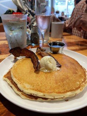 Pancakes topped with soft warm butter and a side of crispy bacon plus warm maple syrup. Cranberry mimosa and glass of water.