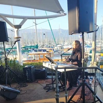 Musica Anima Vocal Facilitator singing while playing the piano with the harbor behind them.