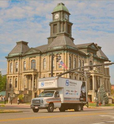 A branded delivery truck for Millersburg Electric is parked in front of a historic courthouse building...