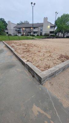 Broken/missing brick on volleyball court. Weeds growing in sand pit.