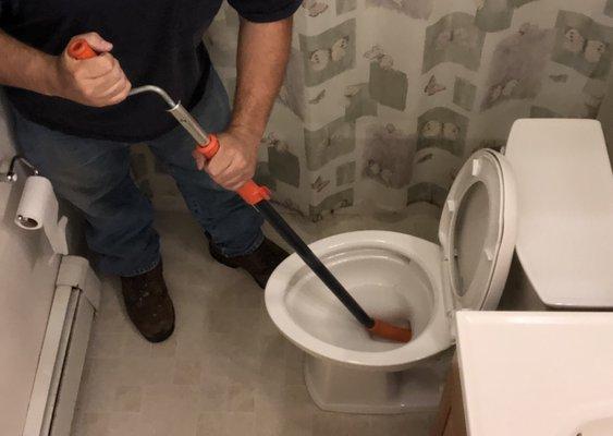 Clear blockage in clogged toilet with auger snake , note protective orange rubber at base of snake to ensure no scratches to porcelain.