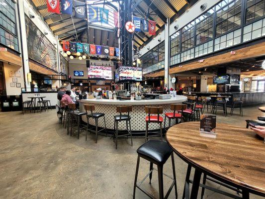 Food court seating area with bar in the center and large screen TVs