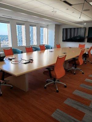 A bright and spacious conference room featuring a long, light-colored wooden table surrounded by modern orange office chairs ...