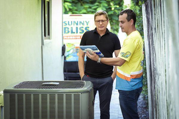 Tech reviewing A/C proposal in front on unit with Sunny Bliss Plumbing & Air van in the background.