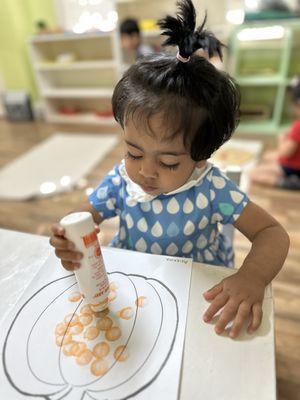 A 20 months old child is making a Halloween art project with teacher's help.