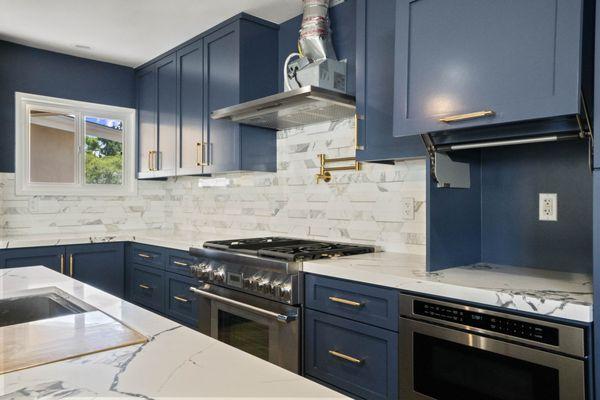 Remodeled kitchen with navy blue cabinets, brass accents, marble countertops, hexagonal backsplash, and high-end stainless steel appliances.