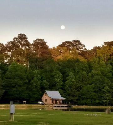 The full moon hangs low over the lake cabin, as the last rays of the sunset grace the tops of the trees.