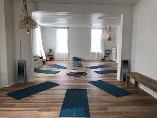 Interior photo of studio showing 8 yoga mats
arranged in an oval shape, natural lighting, room with white walls and white curtains.