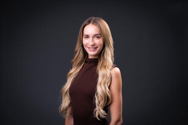 A striking professional headshot of an individual with blonde hair, in an elegant brown dress, and standing against a gray background.