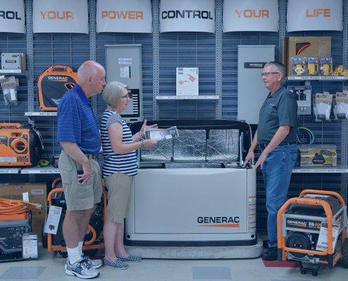 A store showcasing Generac generators. A sales associate is explaining the features of a large home generator to a senior cou...