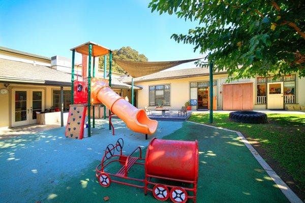 One of our two play structures in the play yard.  Climbing wall, play kitchen underneath, a tube slide.  Classrooms visible in the backdrop.