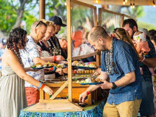 Luau guests gathered around buffet table capturing the lively and communal spirit of a traditional luau at the Voyagers of the Pacific