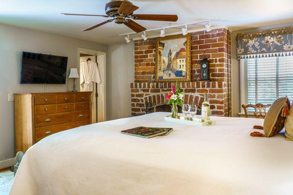 Bedroom with special tray of wine, whine glasses, and floral vase set on tray on soft white bedsheets. A brick fireplaces decorates the wall