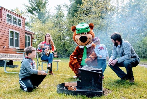 Family posing with Yogi Bear™ while roasting s'mores next to a fire