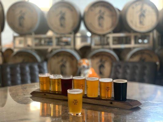 A flight of beer sits on a table with oak barrels in the background.