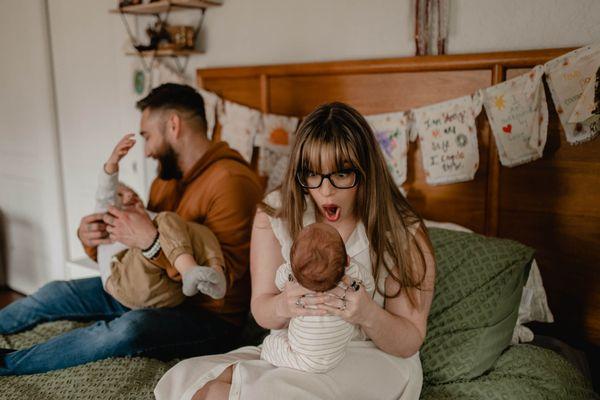 Mom laughs with her newborn baby boy as dad plays with big brother in their master bedroom