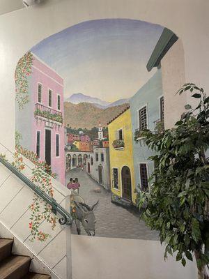 Spanish themed city street mural with a donkey with lovely ladies walking surrounded by pink, yellow and blue buildings.