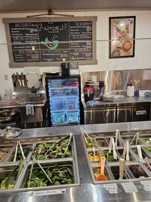 The difference ingredients behind the glass wall to make your own bowl and a menu on the wall in the background.