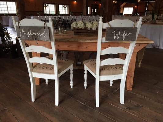 Sweetheart chairs with hanging wooden signs, set up with our six foot farm table, wooden crate, and mercury glass.