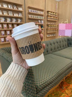 A hand holds a white travel coffee cup with a brown sleeve that says "Grounds" in front of a wall of chocolate bars and a green couch.